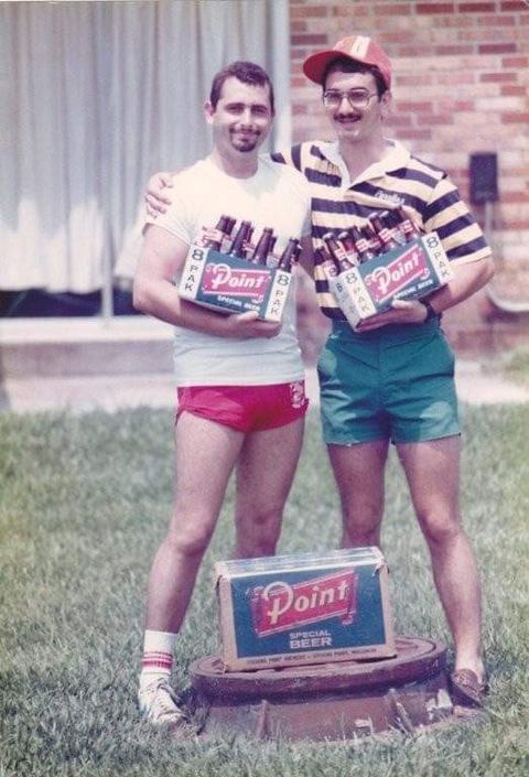 1980's era image show two men outside wearing shorts and t-shirts holding 8-packs of Point Special, with a case of the beer on the ground in front of them.