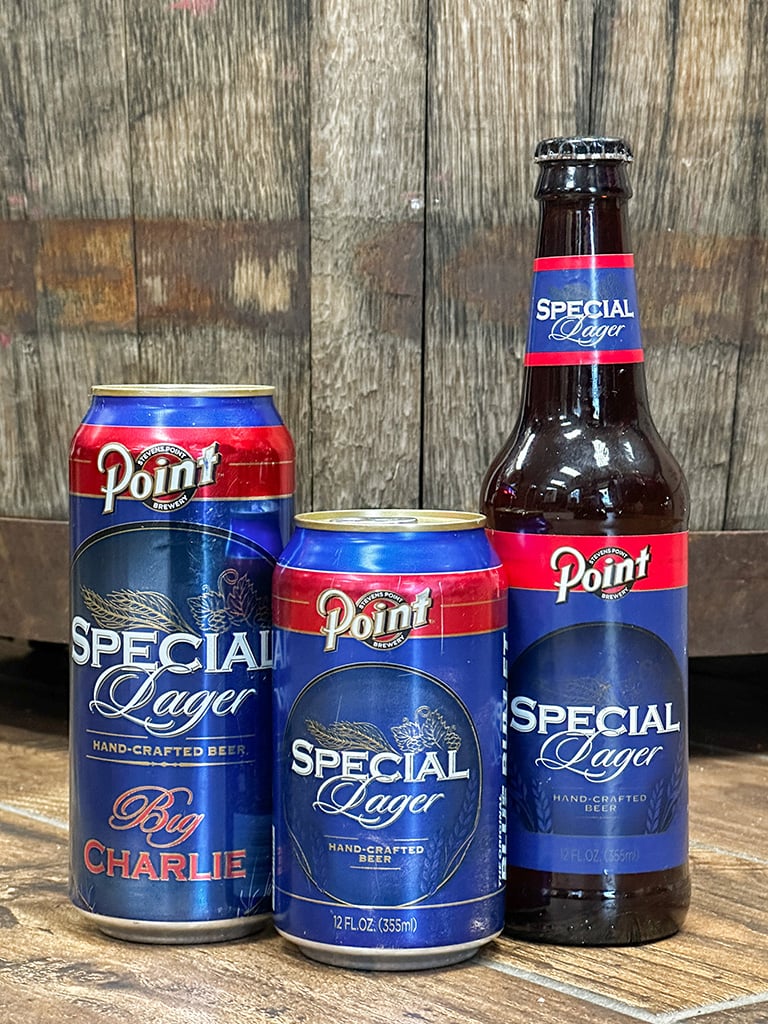 A 16oz can, 12oz can and a bottle of Point Special beer sit in front of a wooden barrel