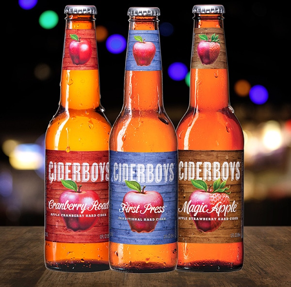 Three bottles of Ciderboys Hard Cider on a wooden table in front of an abstract blurred background of bokeh light